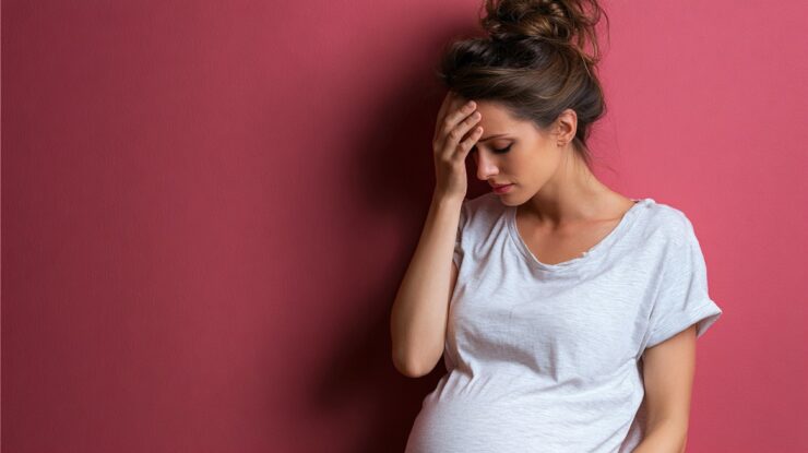 Pregnant woman in a white shirt holding her head, looking stressed against a pink background