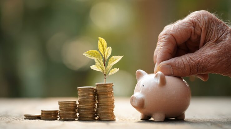 Hand placing a coin into a piggy bank beside stacked coins with a growing plant