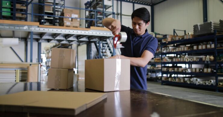 Person sealing cardboard boxes with packing tape on a worktable inside a warehouse environment